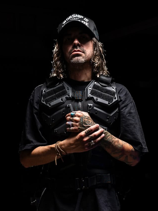 Joel Piper wearing a black cap and vest, with his hands clasped against a dark background